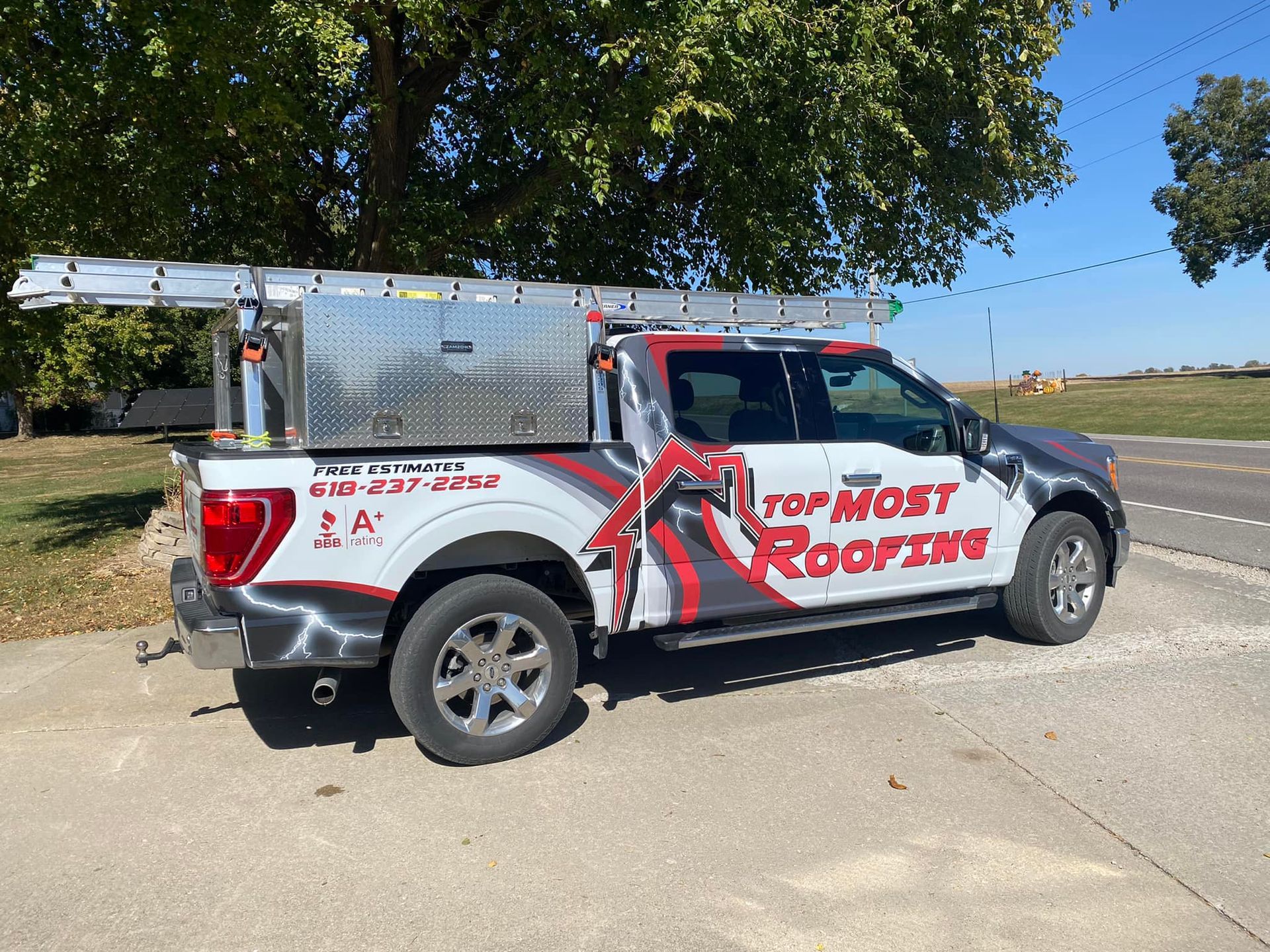 Top Most Roofing truck with ladder rack parked on pavement next to a road.