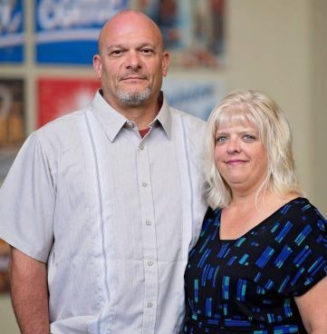 A man in a light grey short-sleeved button-down and a woman in a black patterned top posing together indoors.