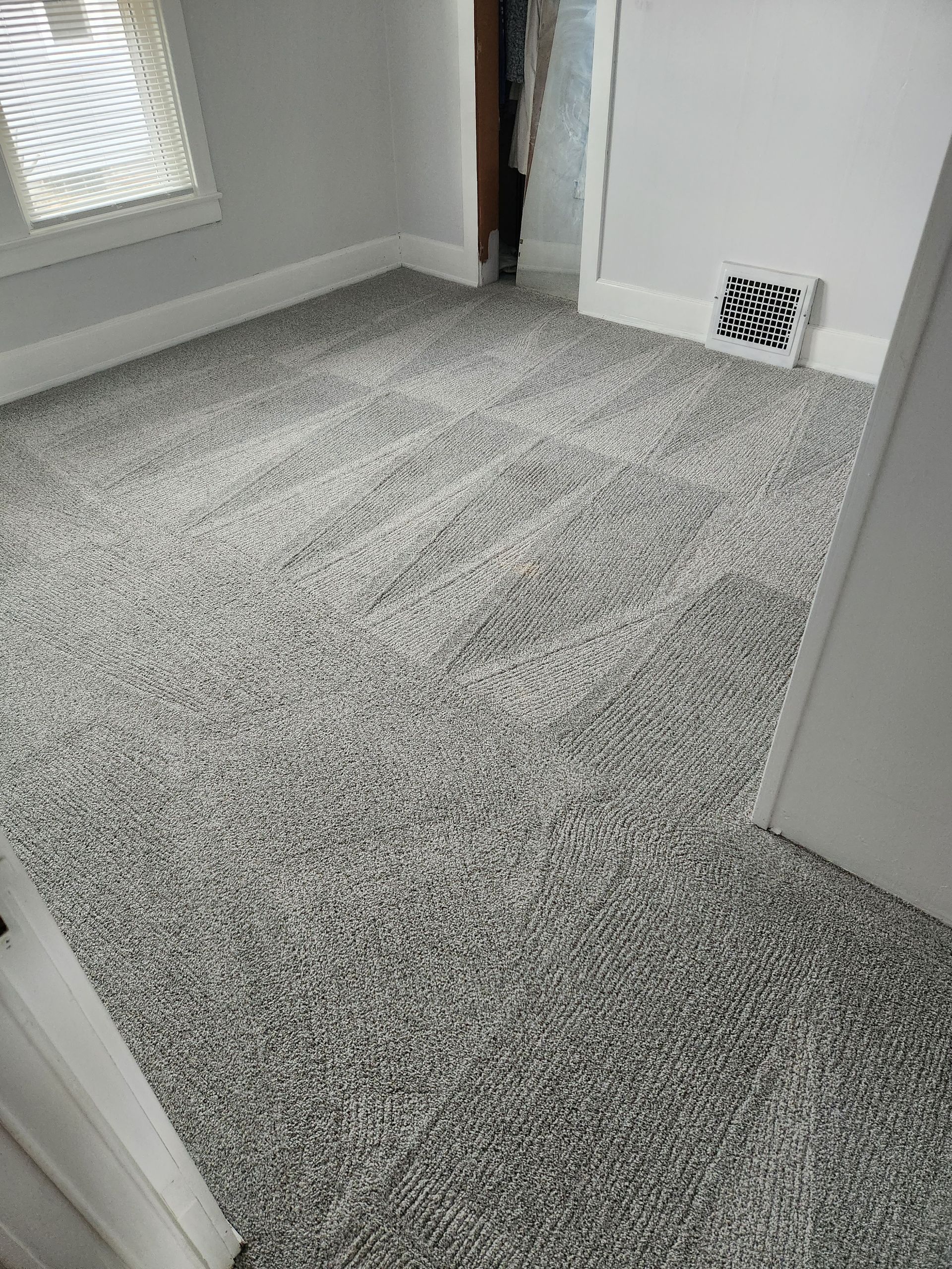 A small, empty room with grey, speckled wall-to-wall carpeting, light grey walls, a window, and a white air vent.