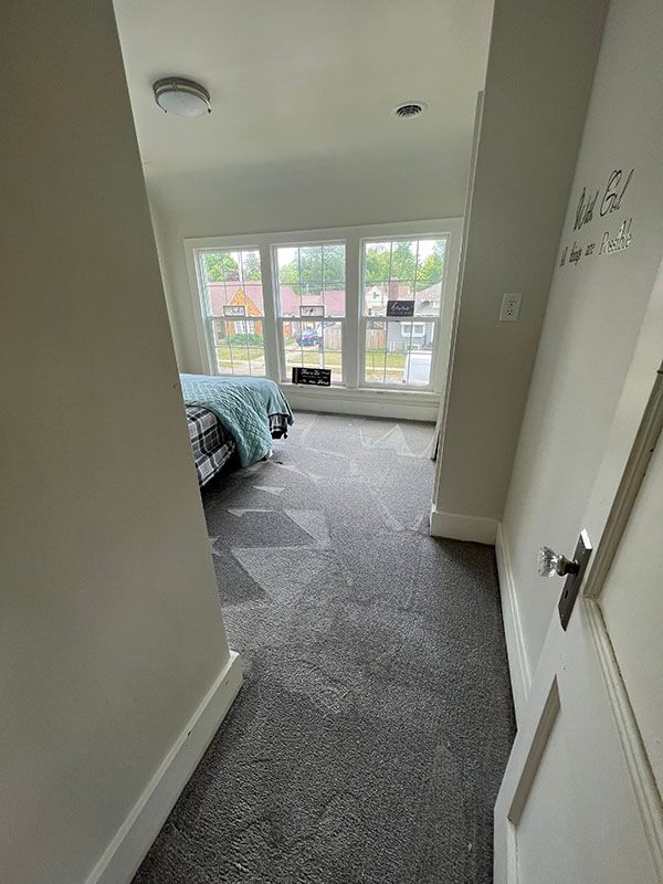 A view from a doorway looking into a bedroom with grey carpet and a bed covered by a light blue blanket near a window.