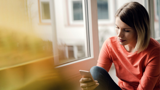 A person in a coral long-sleeved shirt looks down at a smartphone while sitting by a window.