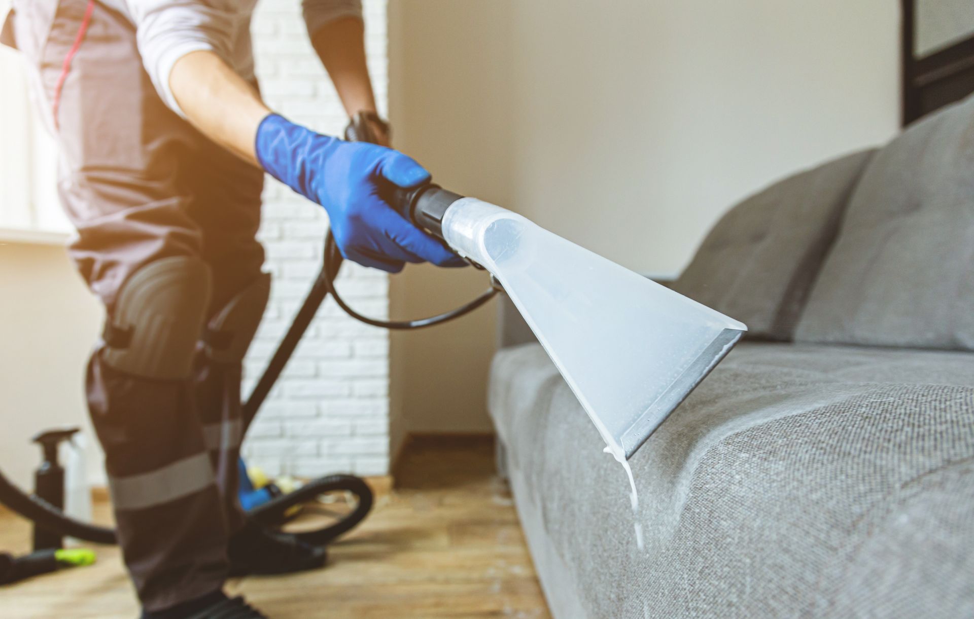 A worker in a uniform and blue gloves uses an upholstery cleaning machine to clean a gray fabric sofa.