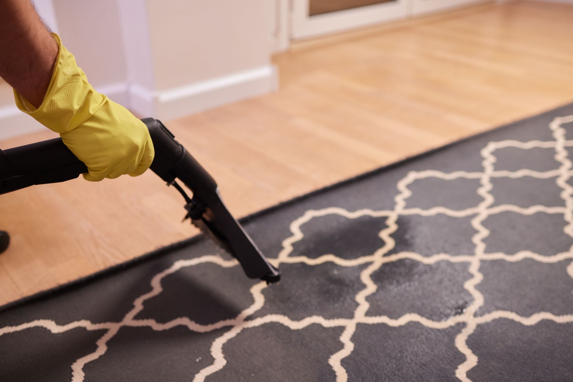 A hand wearing a yellow rubber glove uses a steam cleaner wand to clean a dark gray, patterned area rug.