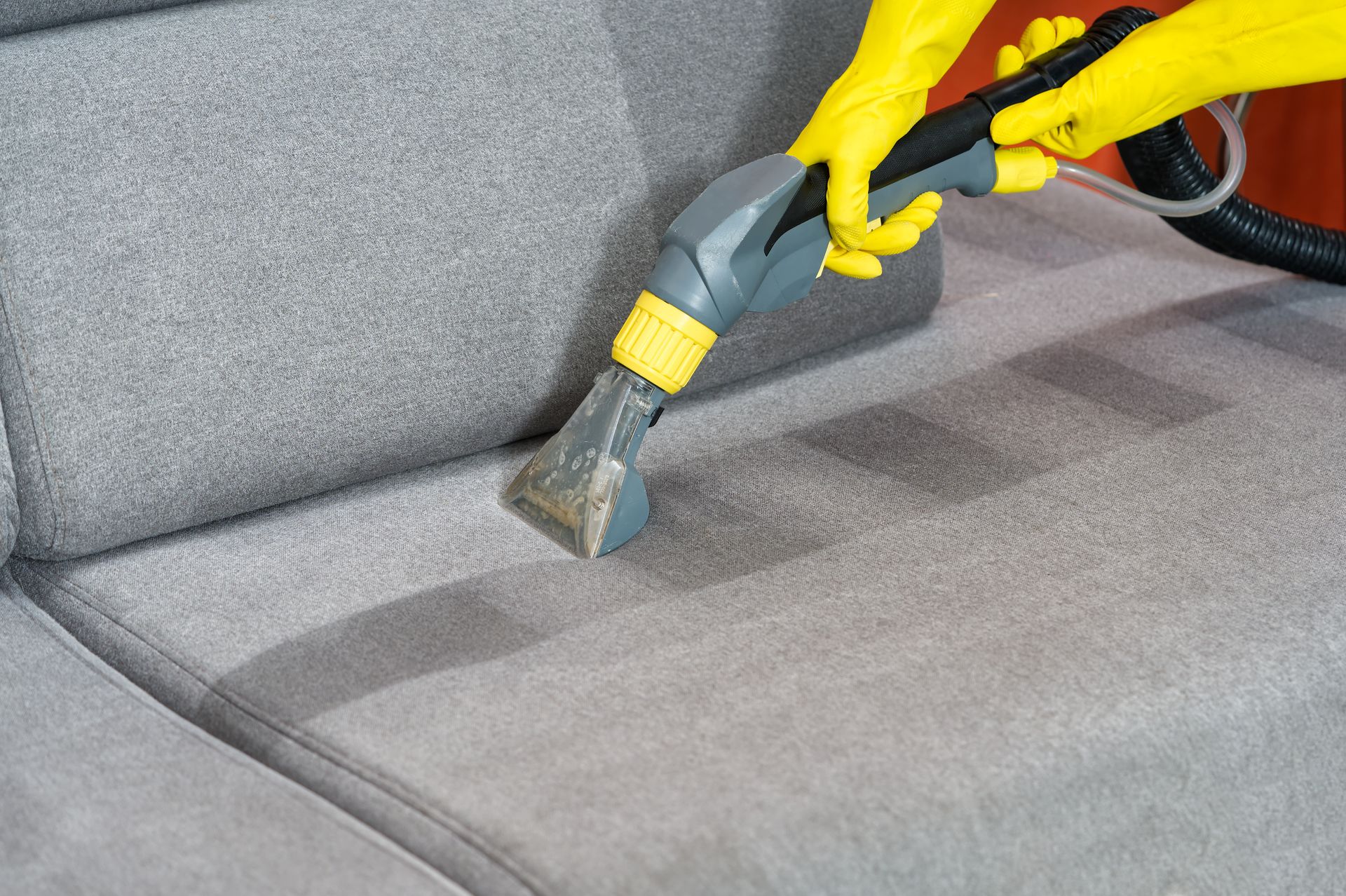 A person wearing yellow rubber gloves uses a machine to deep clean a grey fabric sofa.