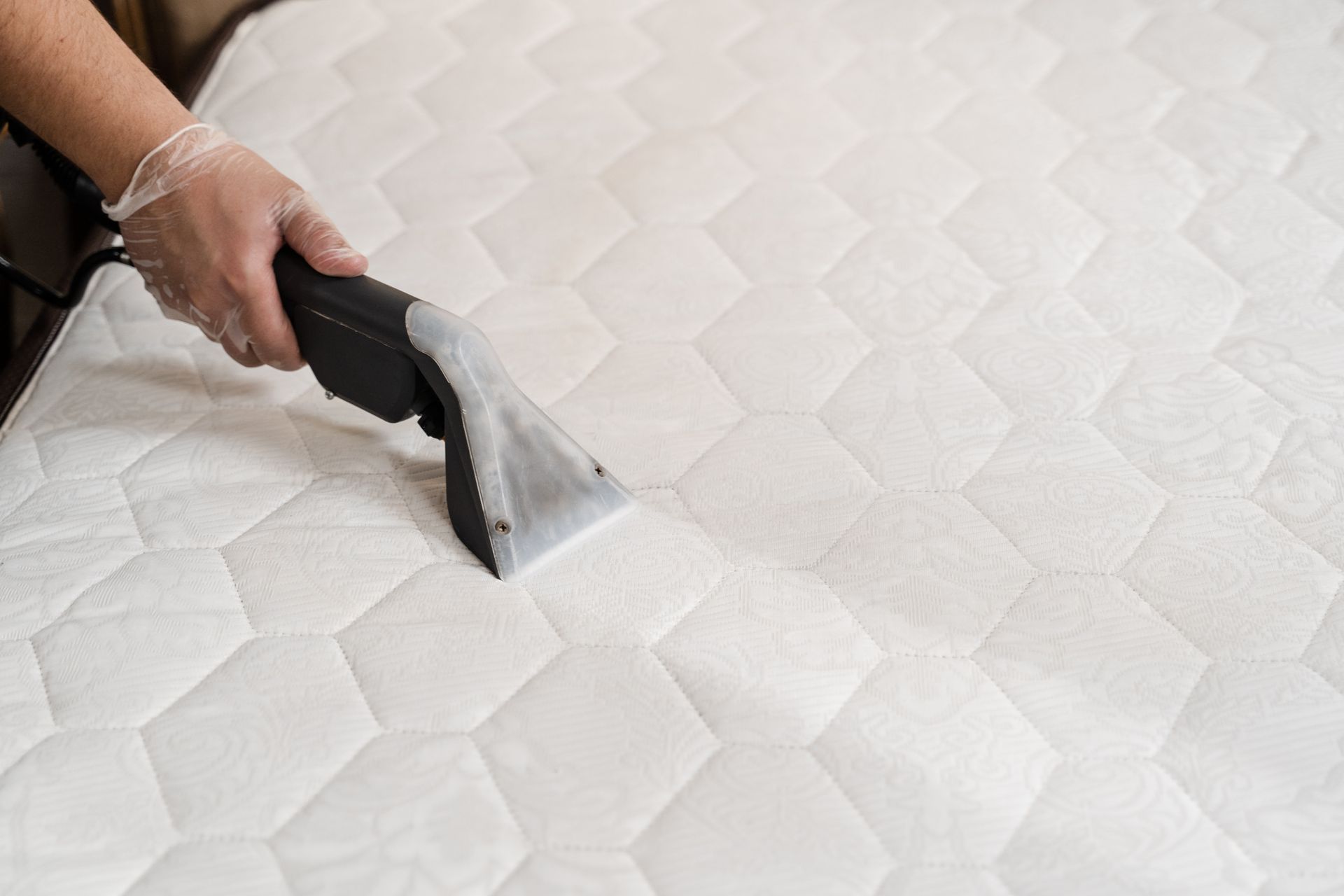 A hand wearing a clear glove uses a steam extraction machine to deep clean a textured white mattress.