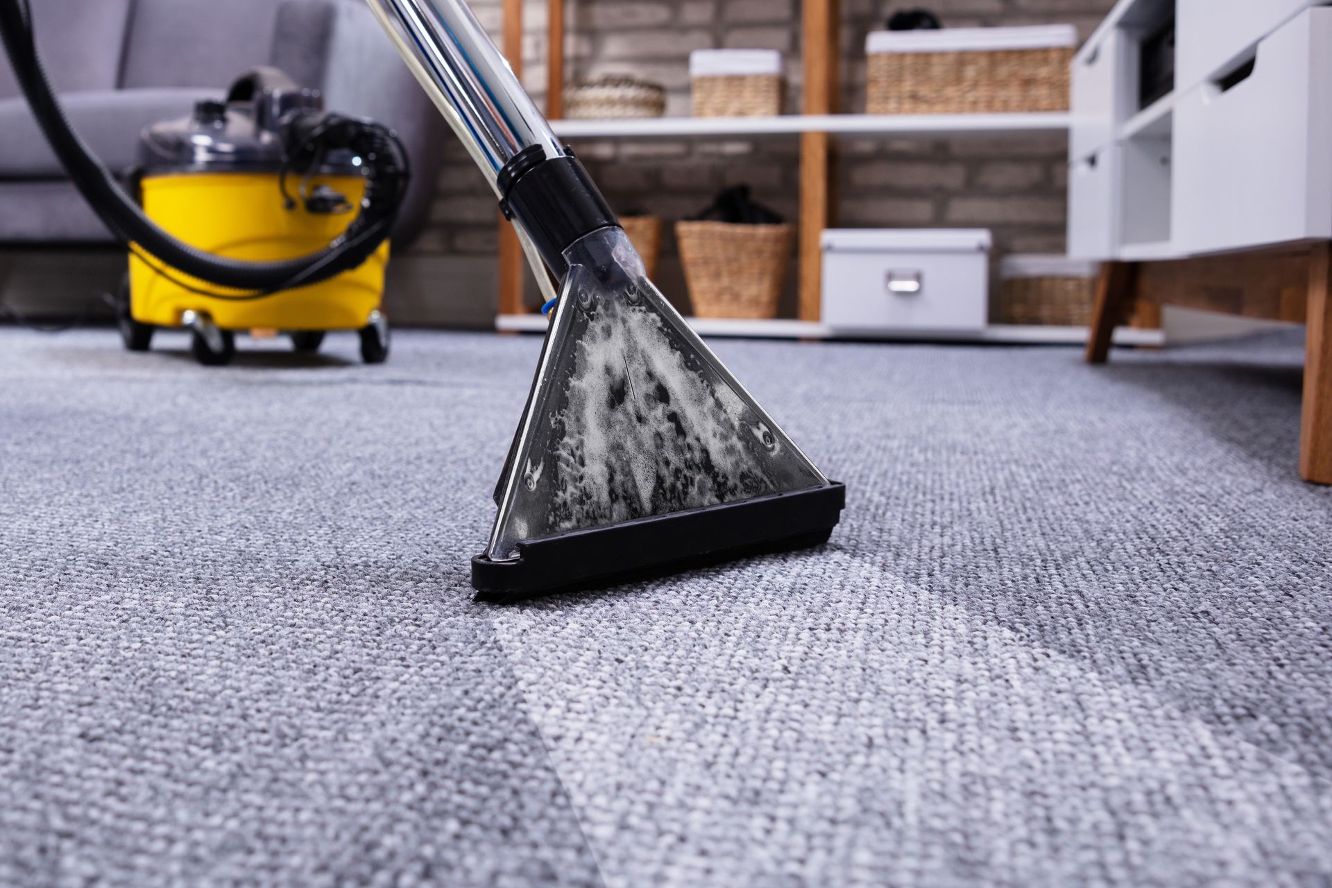 A vacuum cleaner head cleans a gray carpet, showing a distinct line between dirty and clean areas in a living room.
