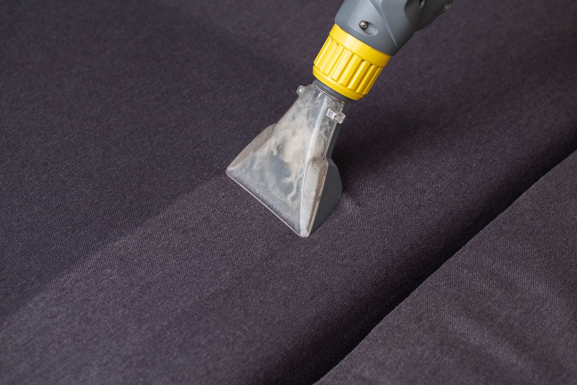 A close-up of a clear upholstery cleaning nozzle extracting dirt from a dark grey fabric sofa.