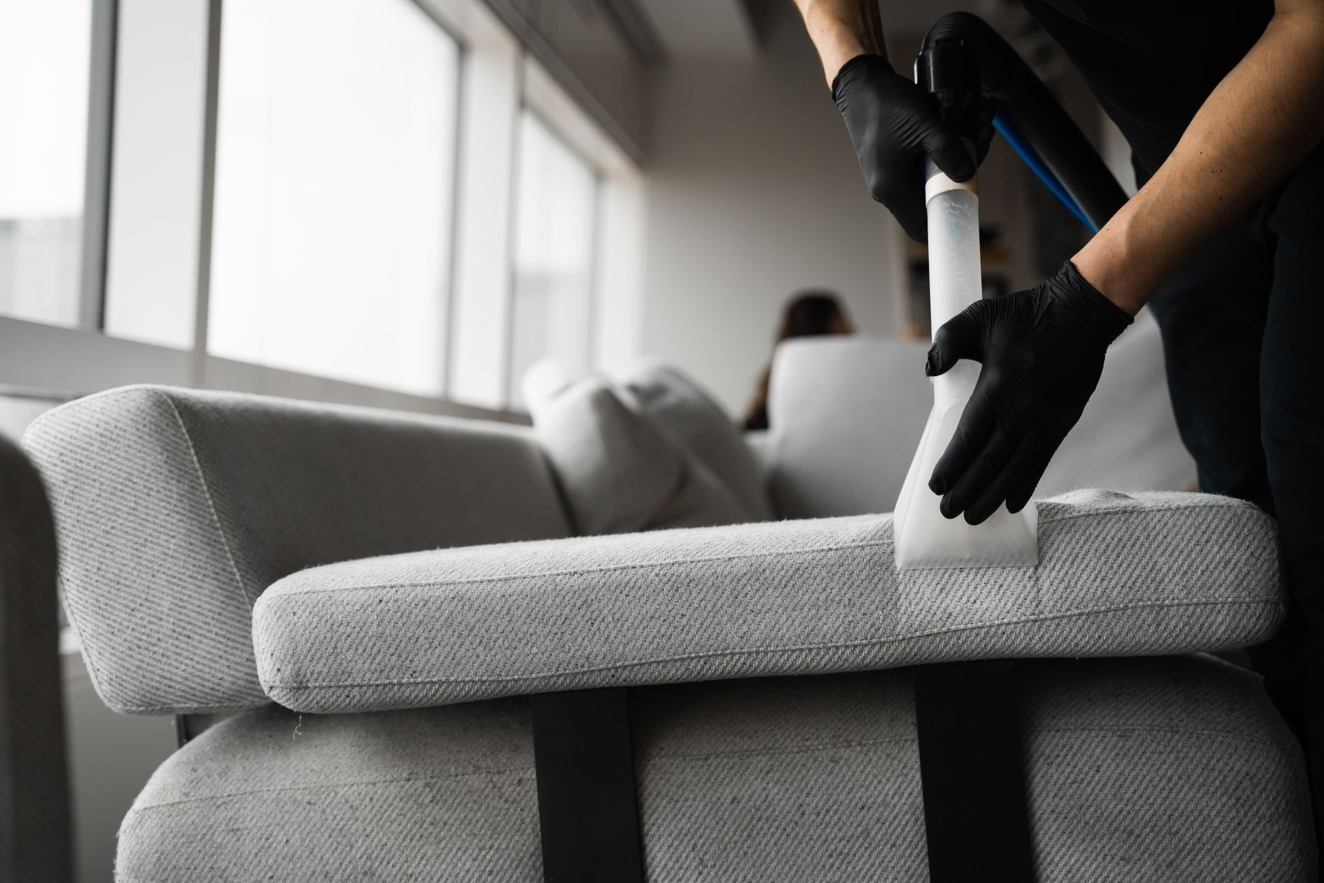 A person wearing black gloves uses a white upholstery cleaning tool to clean a light gray couch.