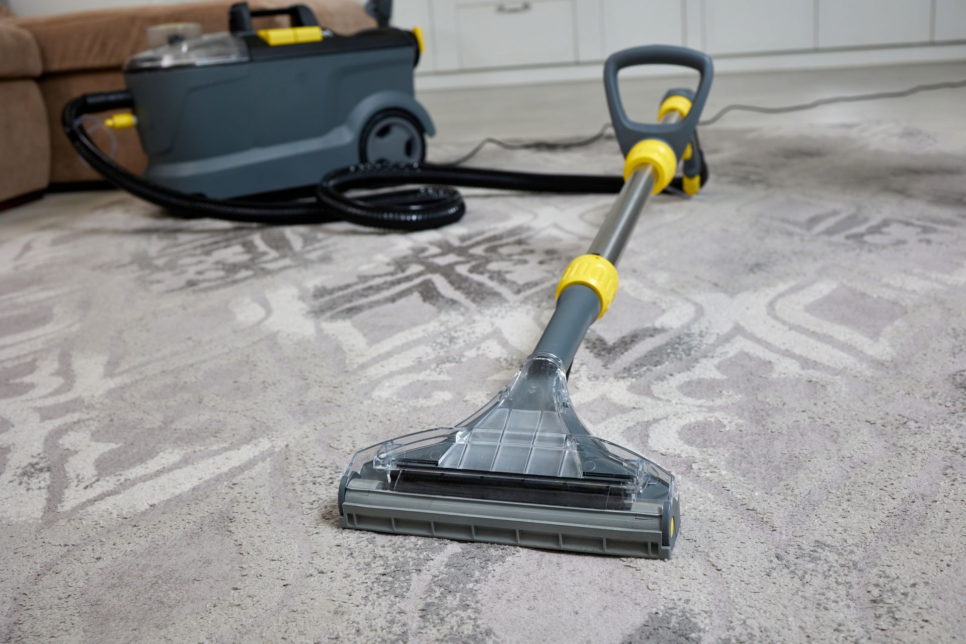 A grey and yellow carpet cleaner nozzle rests on a patterned rug, with the main unit connected by a black hose.