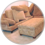 A tan fabric sofa with several decorative pillows and a matching patterned rectangular ottoman on a light carpet.