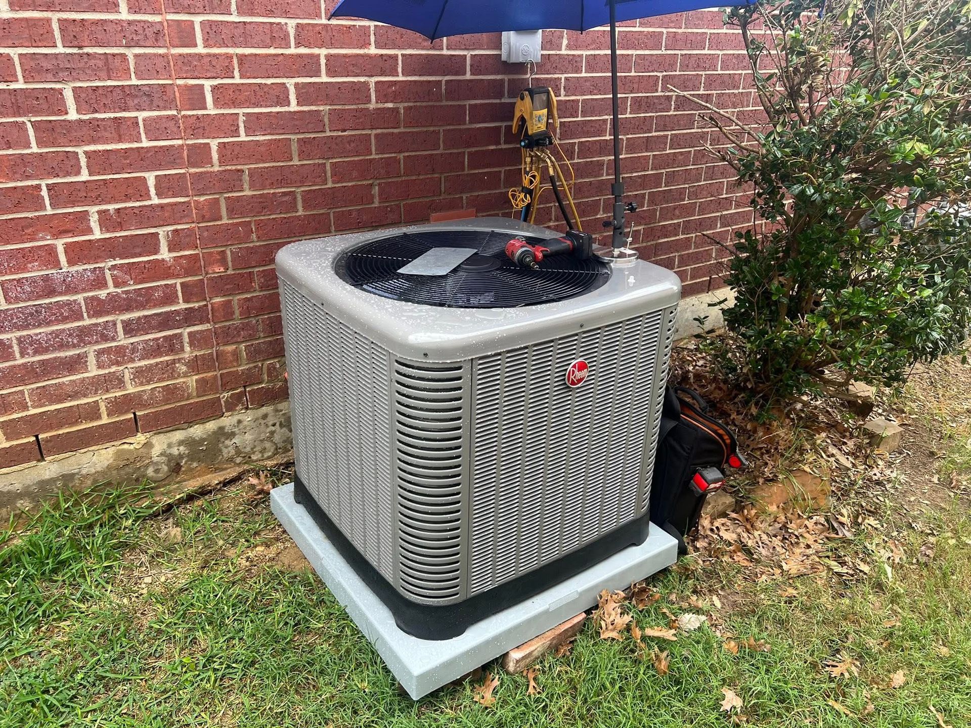 Outdoor air conditioner unit on a concrete pad beside a brick wall and bushes, under a blue umbrella.