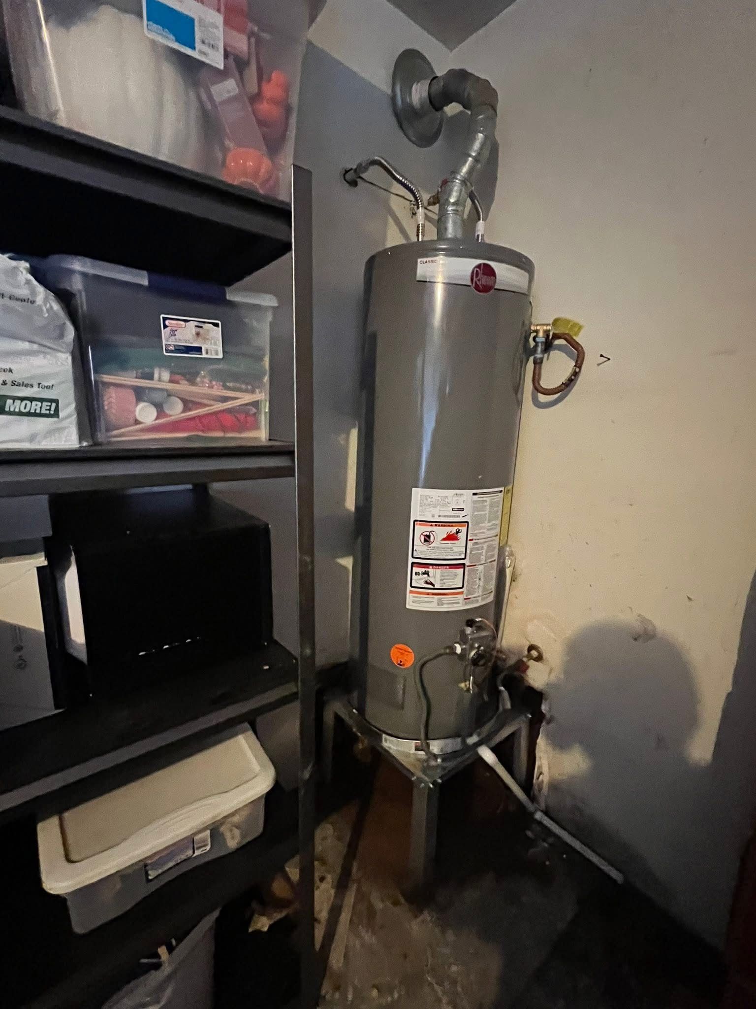 Tall gray water heater with pipes in a utility closet beside black shelving and storage bins