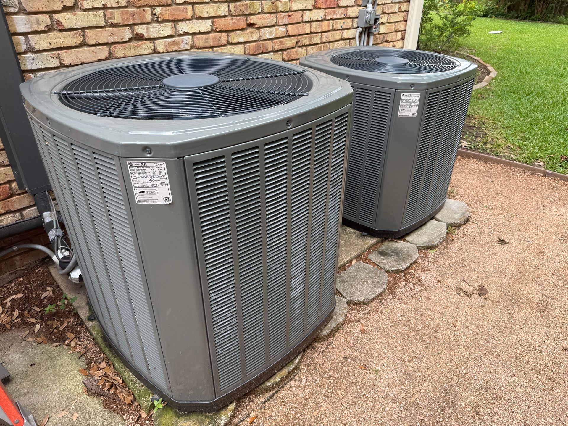 Two outdoor air conditioning units beside a brick wall on a gravel and stone pad.