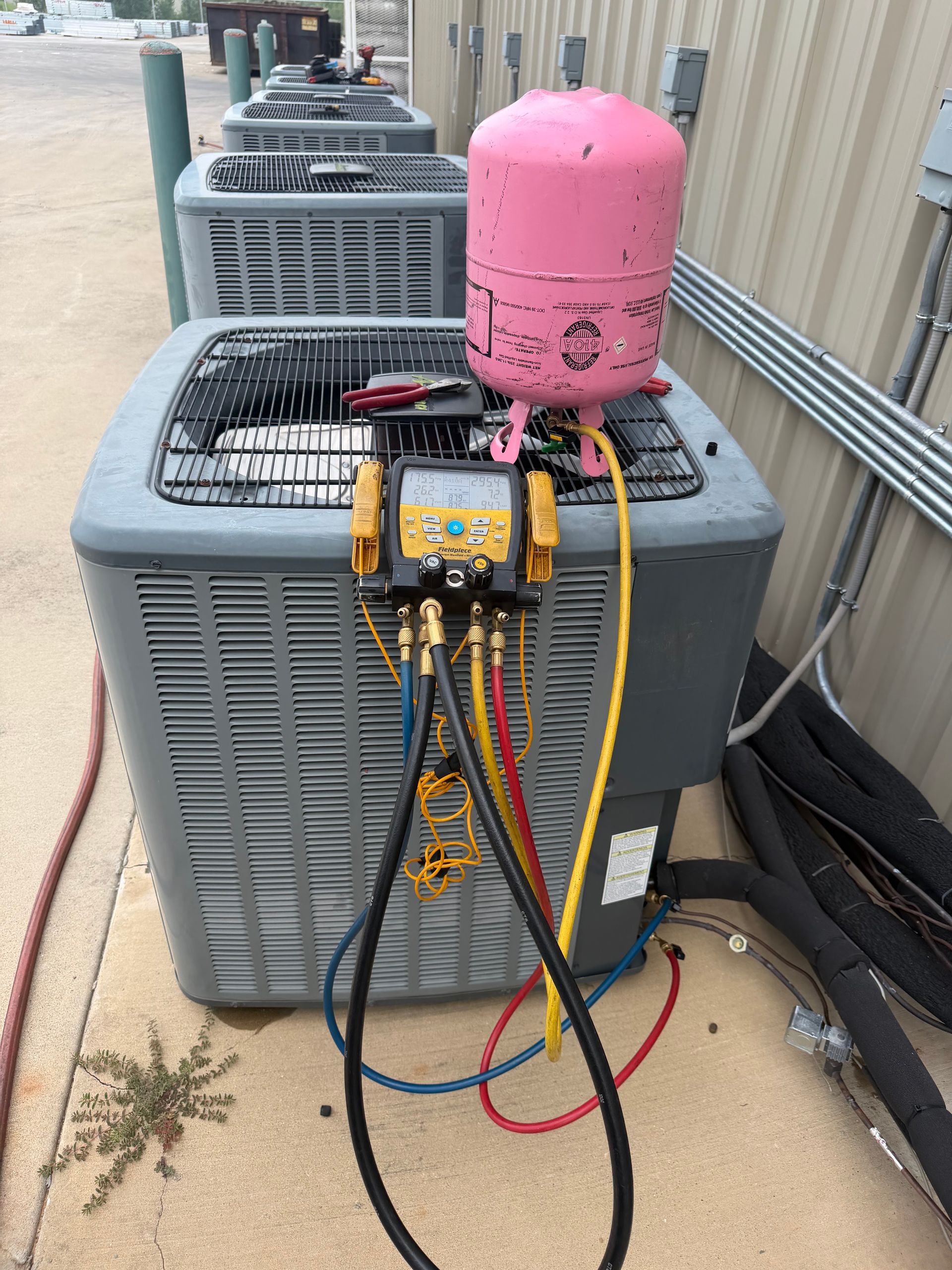 Outdoor HVAC unit with pink compressor tank and attached gauges, hoses, and wiring on the side of a building