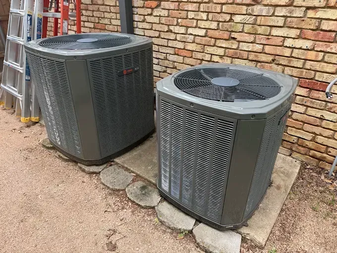 Two outdoor air conditioning units beside a brick wall on concrete pads.
