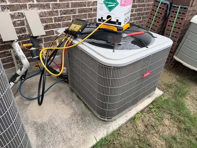 Outdoor HVAC unit with gauges and hoses on top, beside a brick house and grassy yard