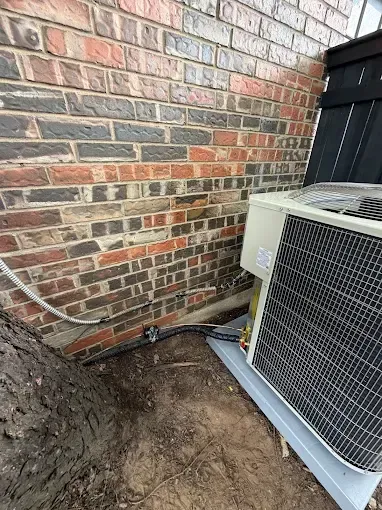 Outdoor HVAC unit beside a brick wall, with exposed dirt and pipes near the foundation.