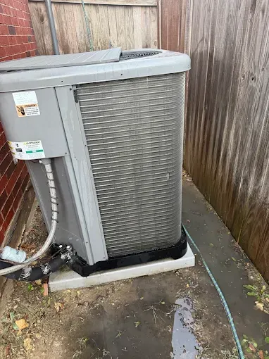 Outdoor air-conditioning unit beside a wooden fence, with water puddled on the ground.