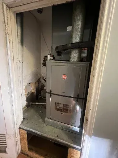 Residential furnace in a closet-like utility space with exposed ductwork and a small step opening.