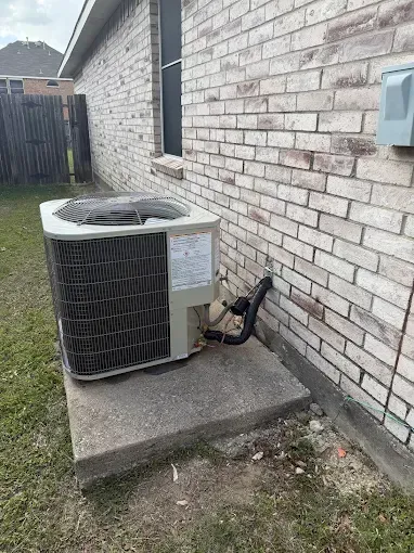 Outdoor air conditioning unit on a concrete pad beside a brick house, with exposed pipes and wiring.