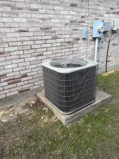 Outdoor air conditioner unit beside a brick wall, with utility boxes mounted nearby on a concrete pad.