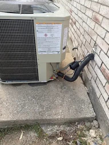 Outdoor air conditioning unit beside a brick wall on a concrete pad with pipes and wiring exposed.