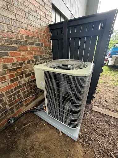 Outdoor air conditioning unit beside a brick house wall and black fence.
