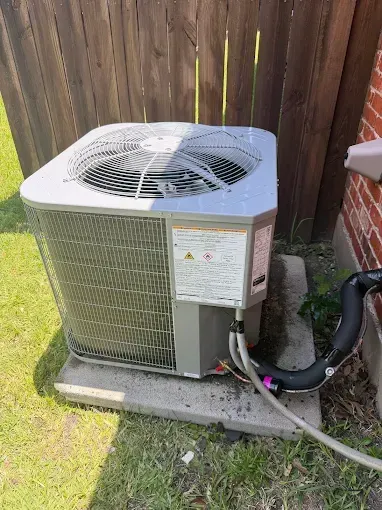 Outdoor central air conditioning unit beside a wooden fence on a concrete pad, with connected hoses.