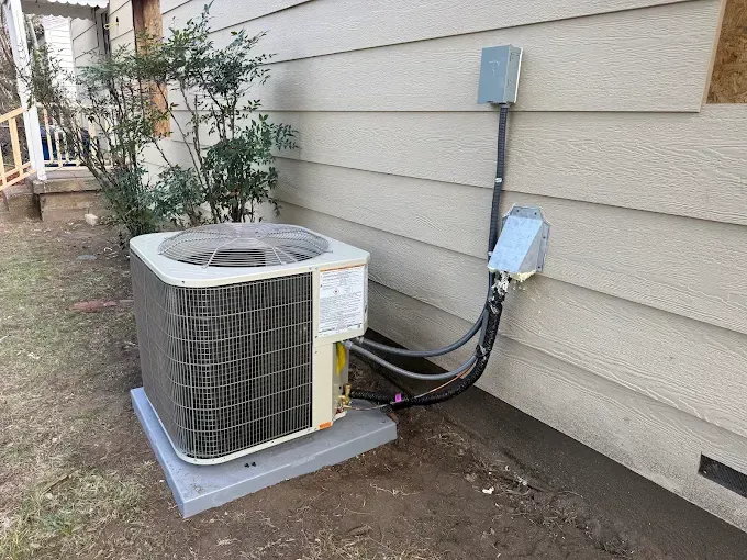 Outdoor air conditioning unit beside a house, with conduit and electrical boxes on the siding.