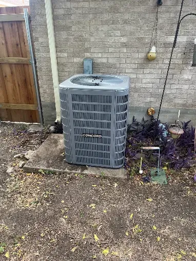 Outdoor HVAC condenser unit beside a brick wall and wooden fence, with mulch and garden debris nearby