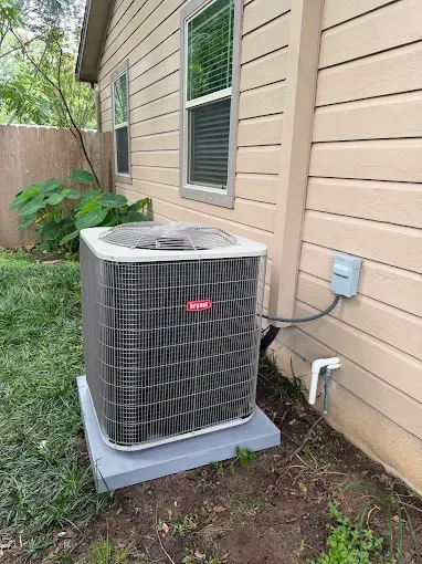 Outdoor air conditioning unit beside a beige house with a window and utility lines nearby