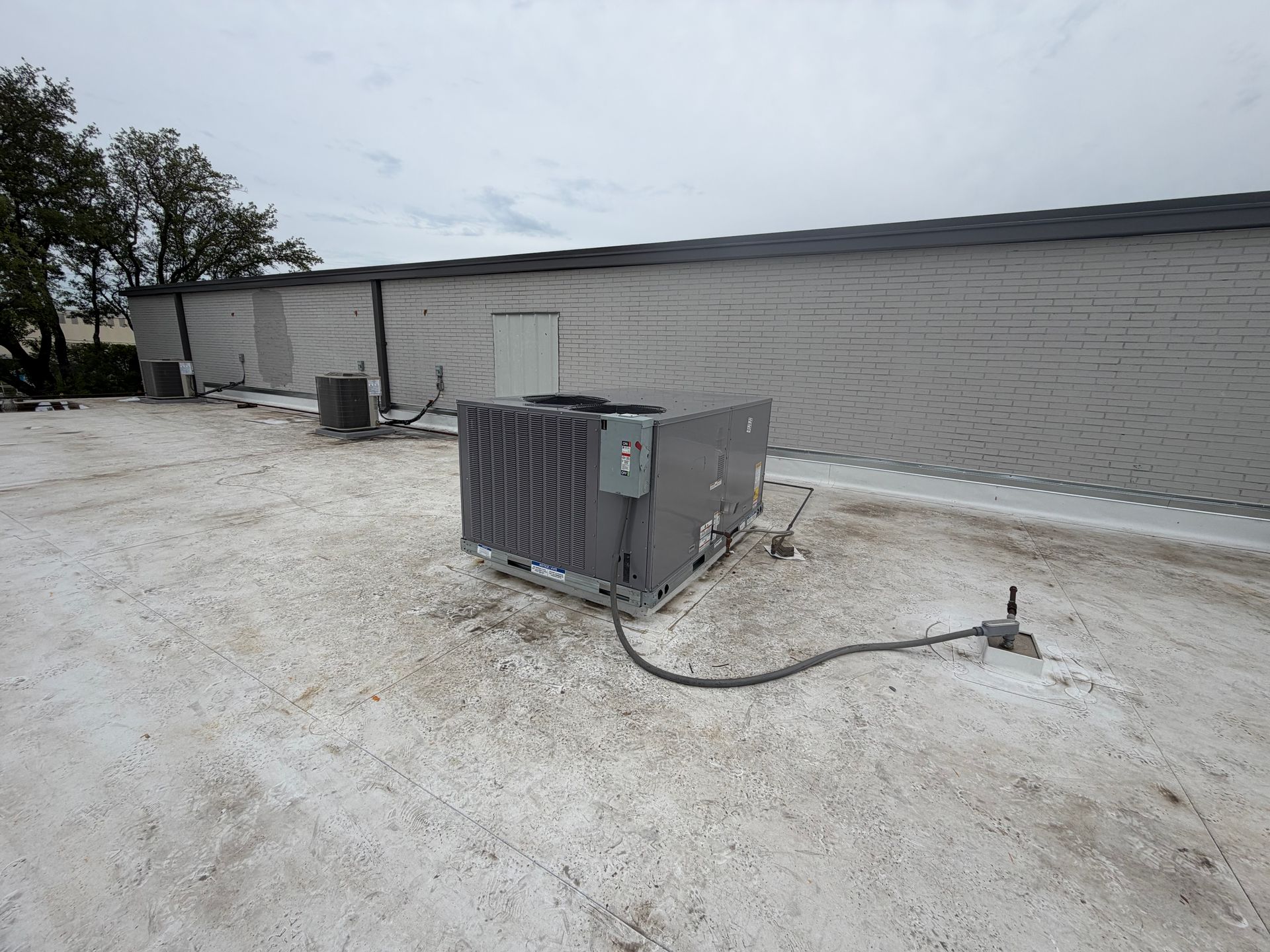 Outdoor HVAC unit beside a long gray building on a gravel lot, with trees in the background