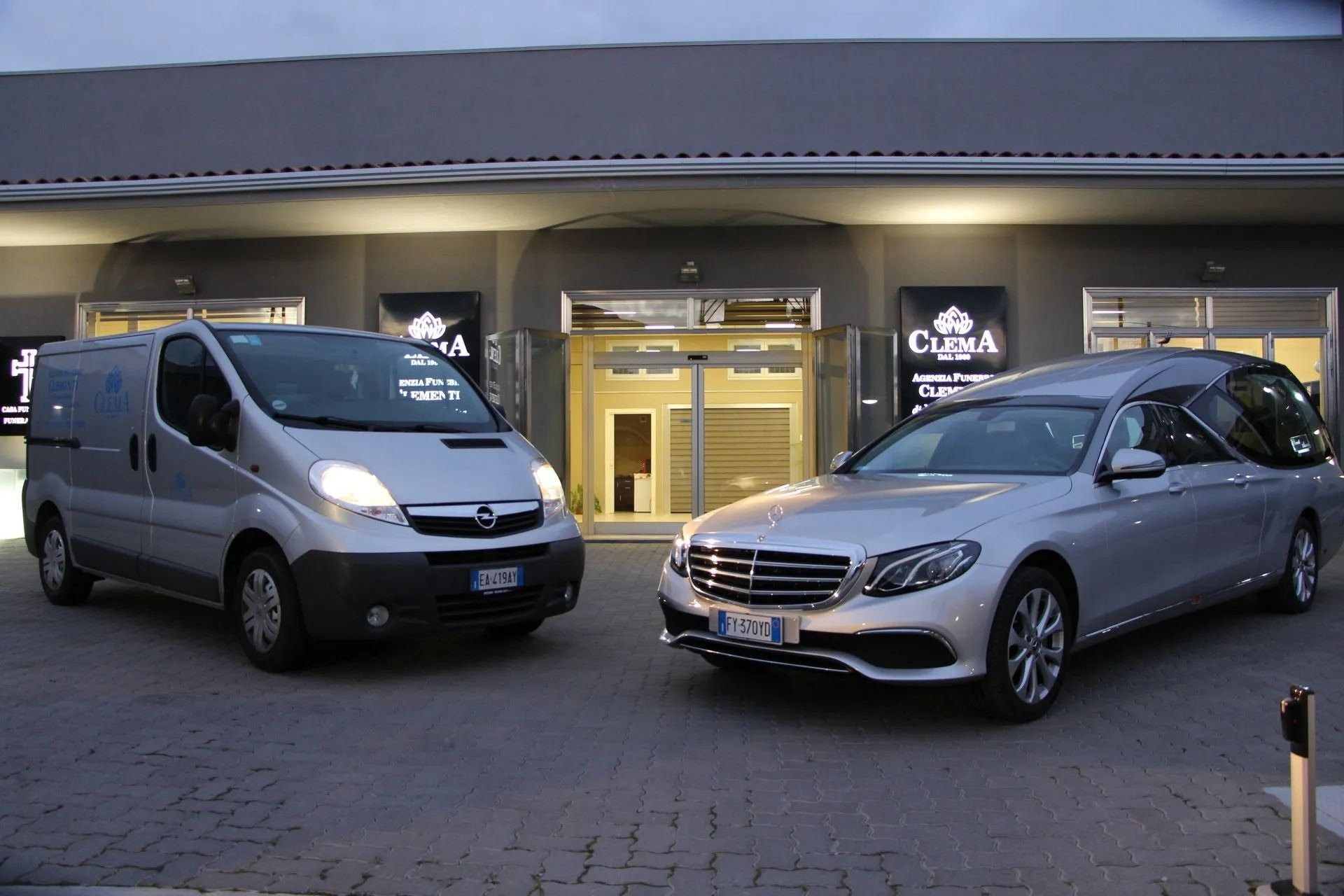 Silver car and white van parked outside a building at dusk, near a lit entrance.