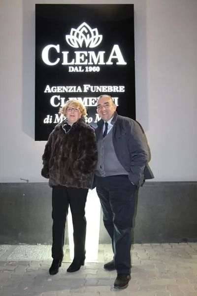 Two people stand arm in arm outside a funeral agency sign reading “Clema.”