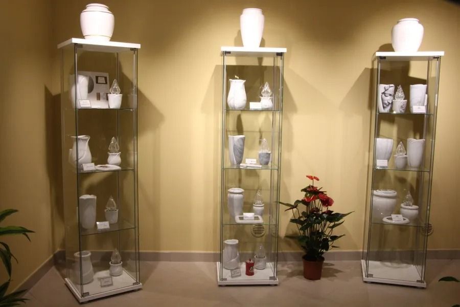 Three glass display cabinets with white ceramic items on shelves in a beige room, with a potted plant centered below.