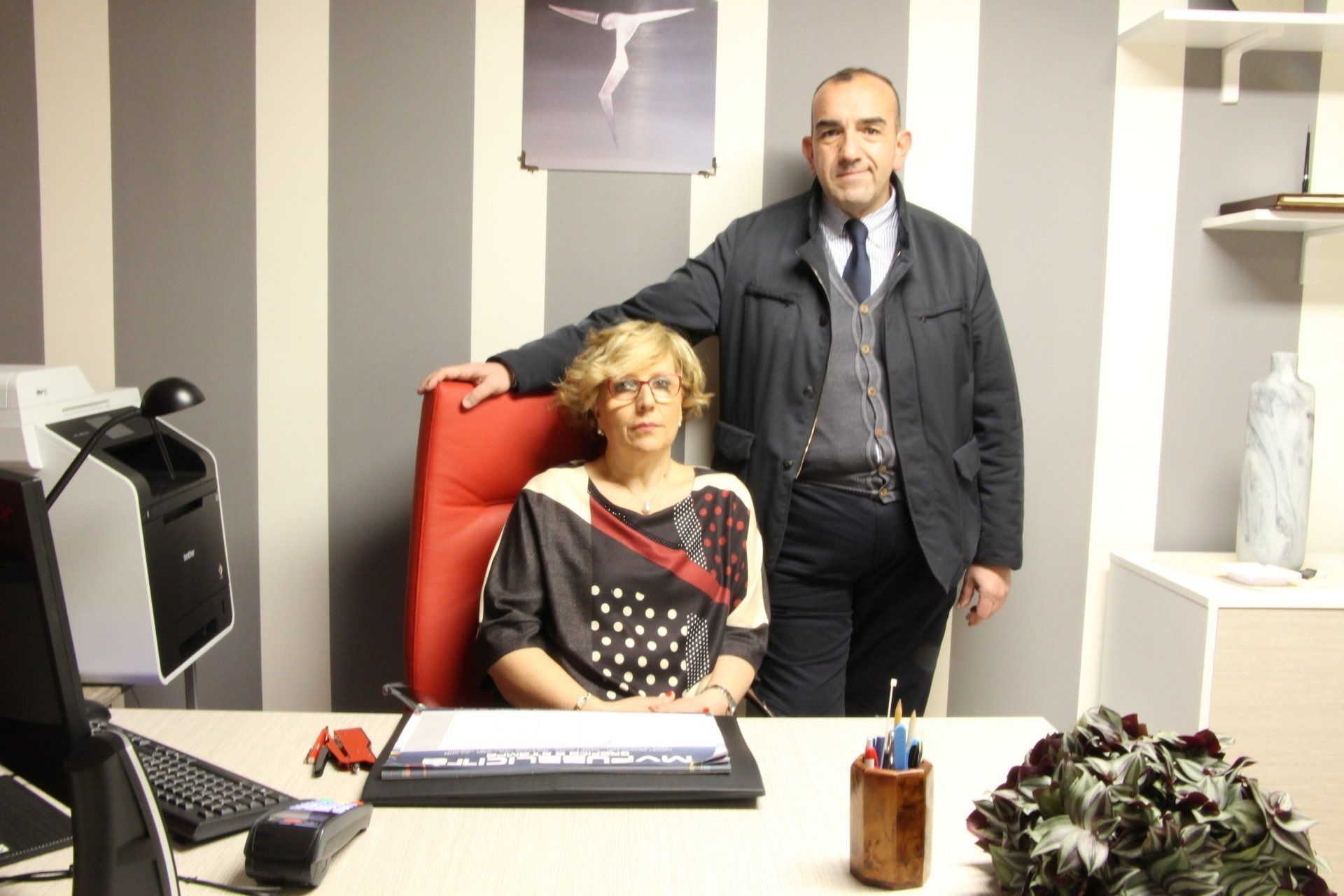 Two people in an office, one seated at a desk and one standing beside a red chair