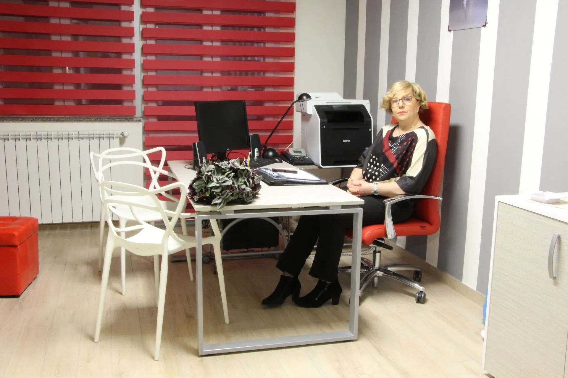 Woman seated at a desk in a modern office with computers, white chairs, and red blinds