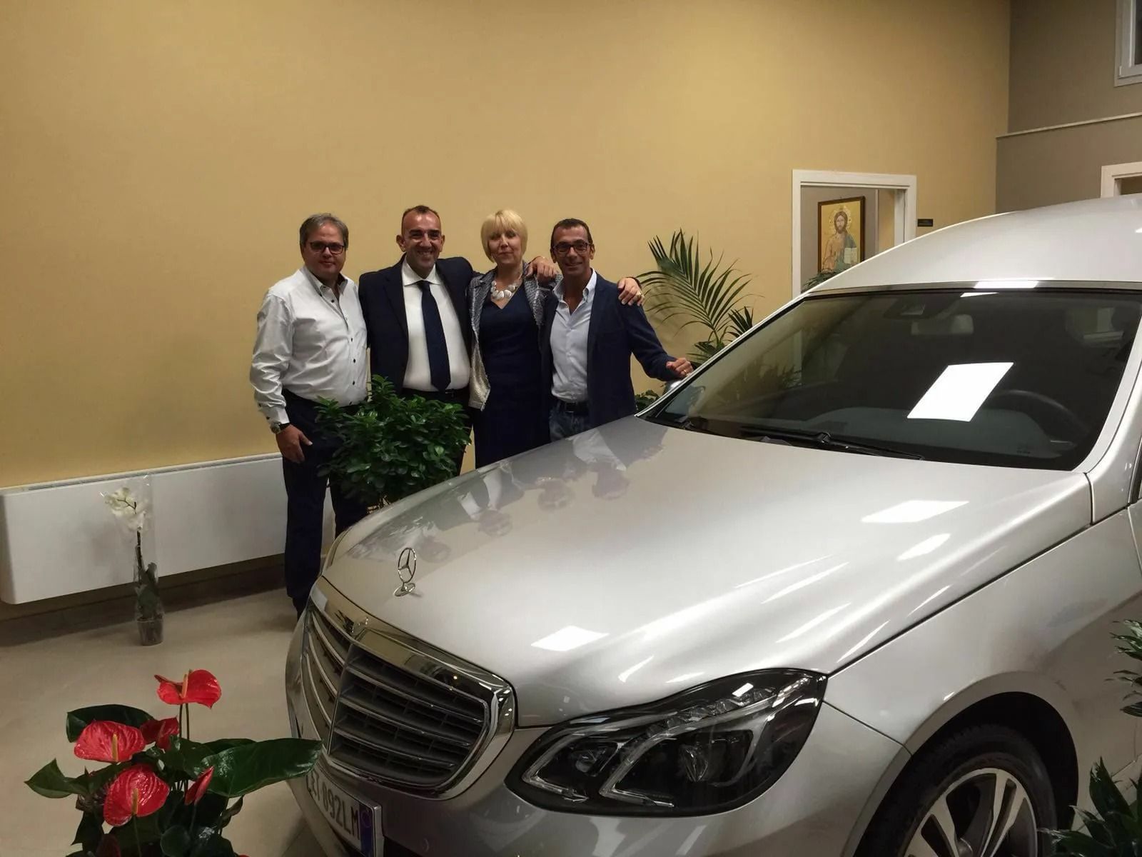 Four men stand beside a silver Mercedes in a lobby, with a potted plant and flowers in the foreground.