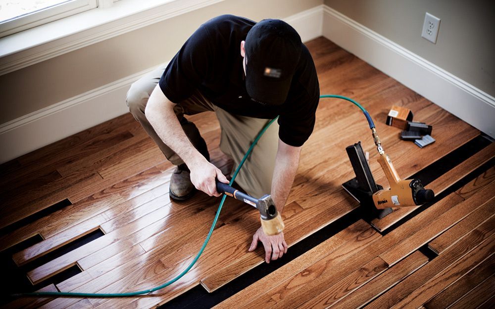 Person installing hardwood flooring with tools and green hose in a room with unfinished floor boards