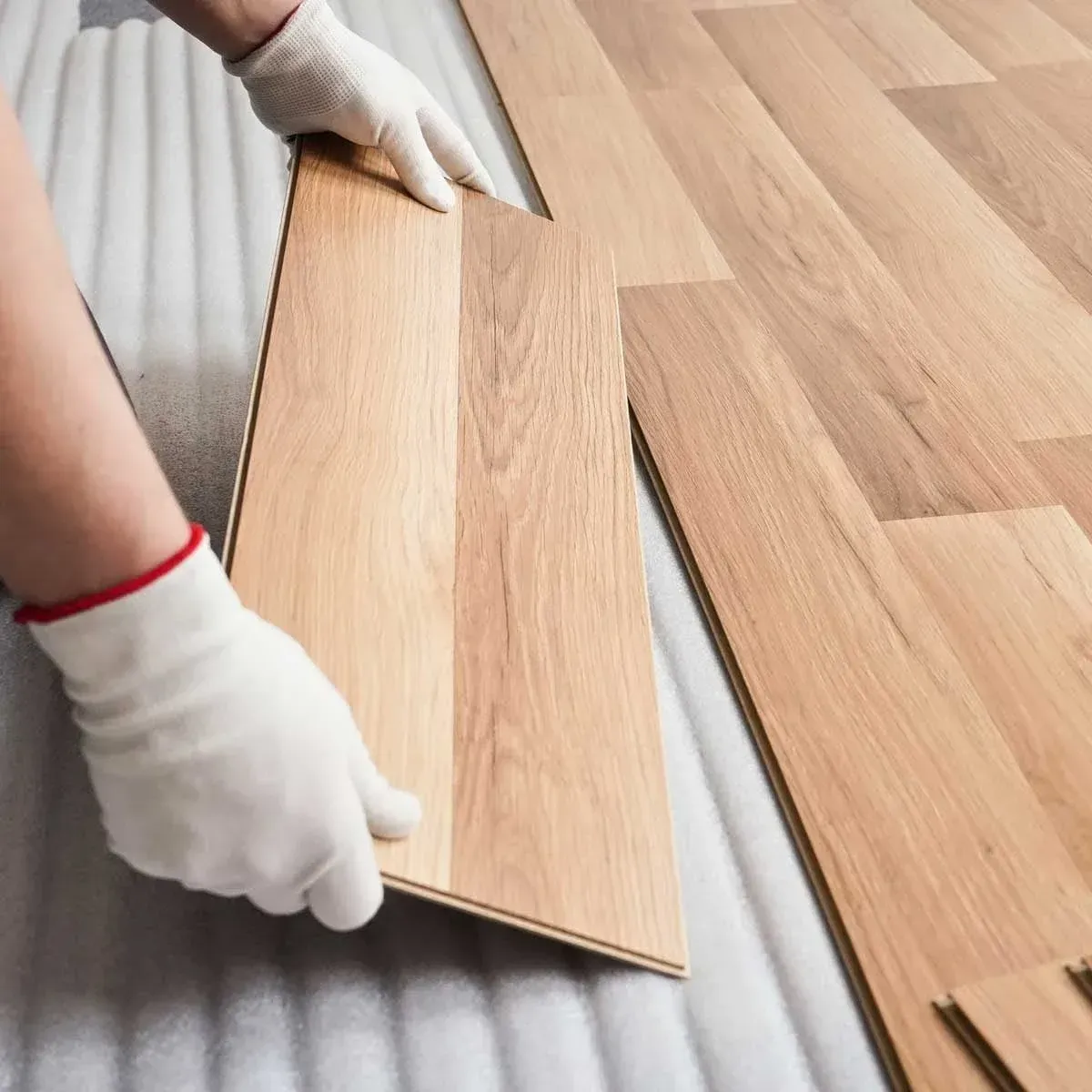Gloved hands fitting a light wood floor plank into place on a hardwood floor