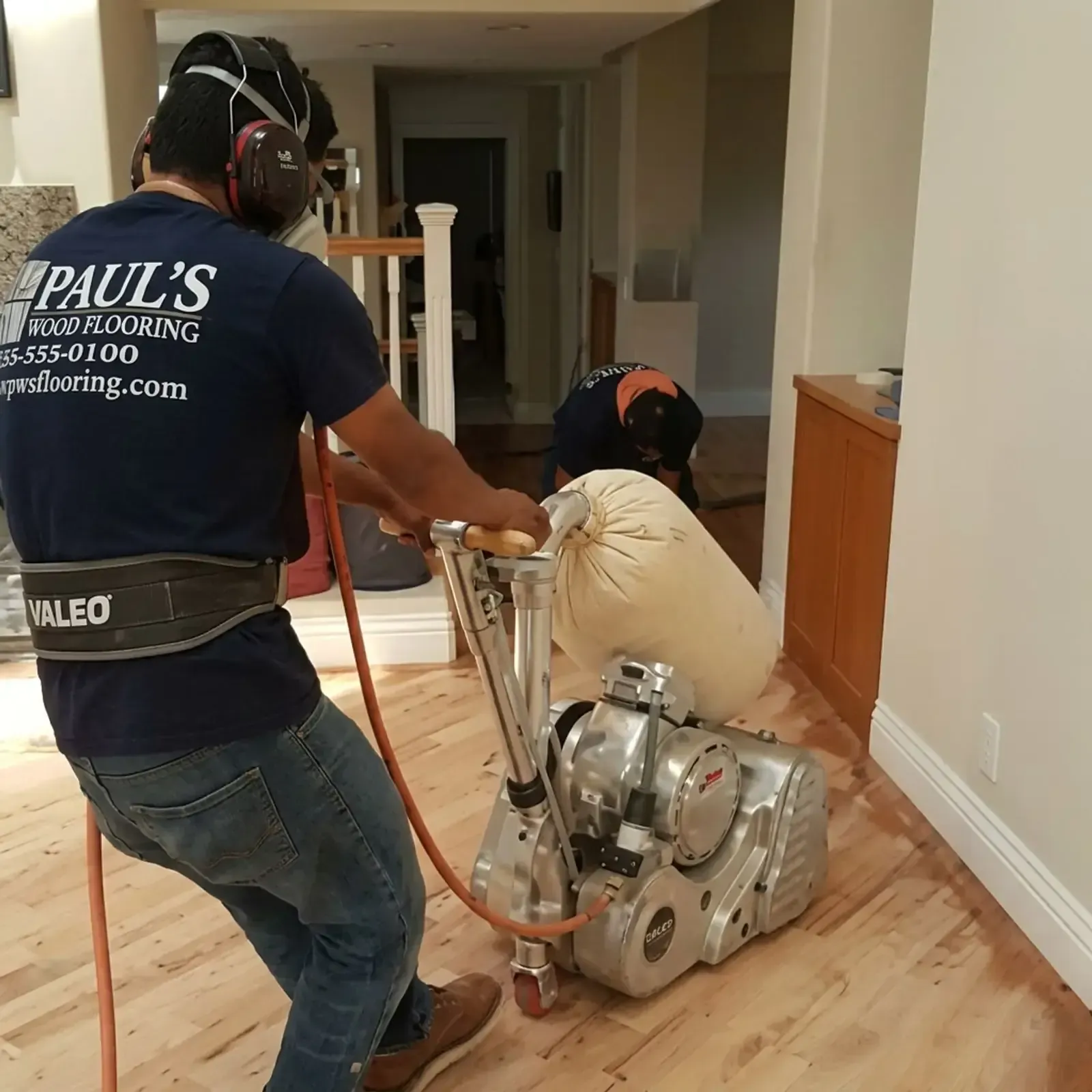 Two workers moving a floor sanding machine across a hardwood hallway inside a home