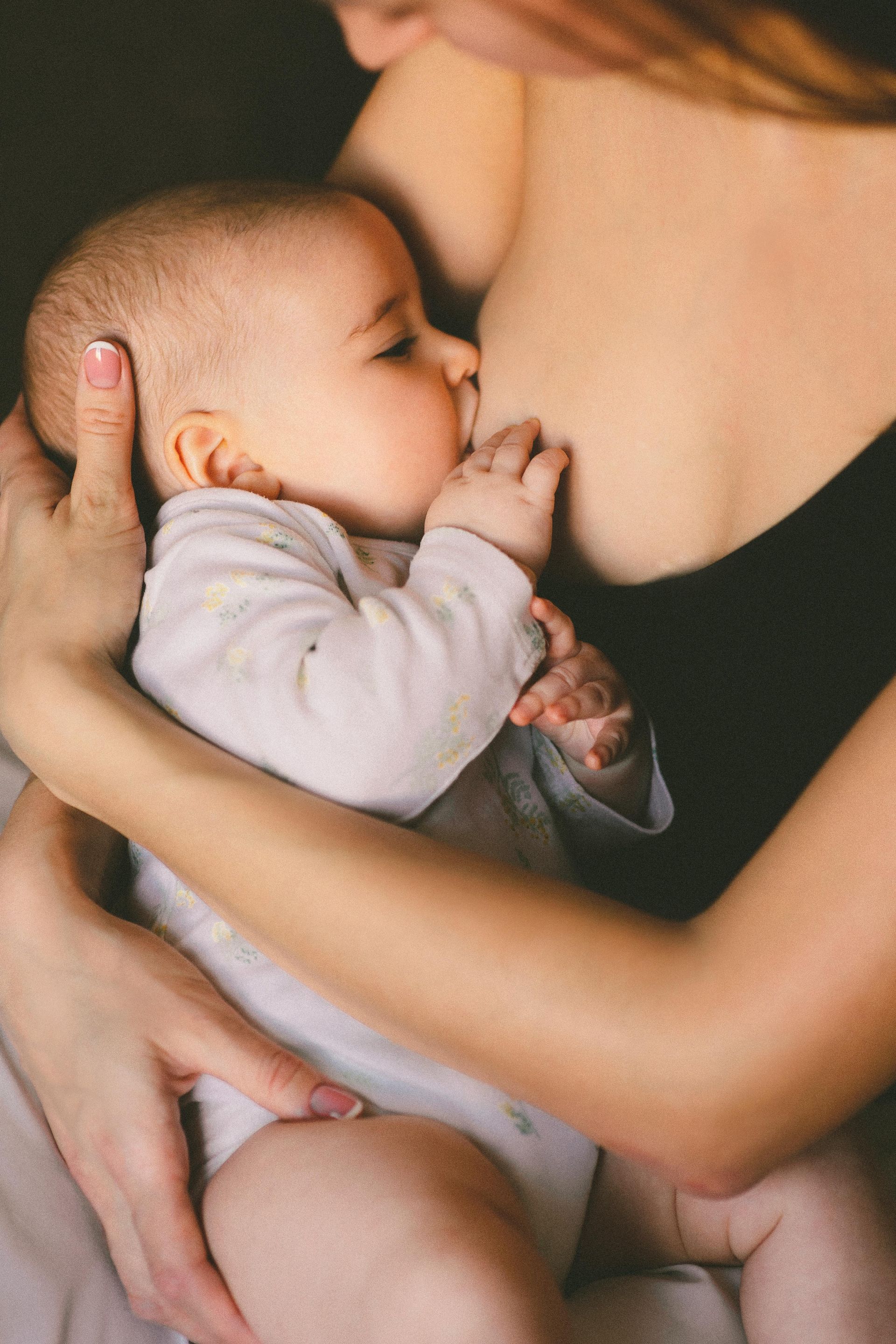 Baby nursing from parent in a dark setting, parent's arm supporting the baby, baby's hand near face.