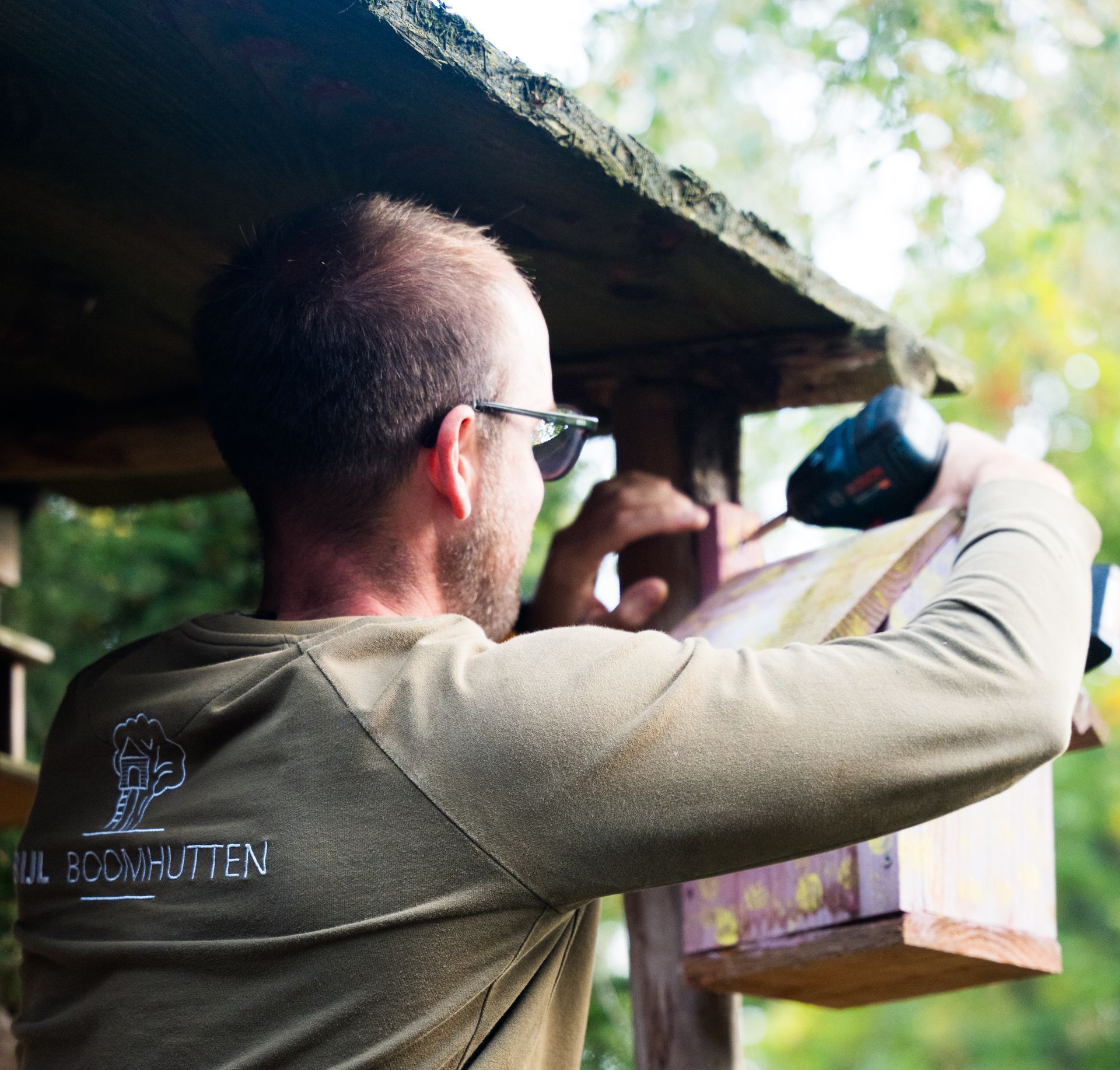 Een man gebruikt een boor om een ​​vogelhuisje aan een houten constructie in de tuin te bevestigen.