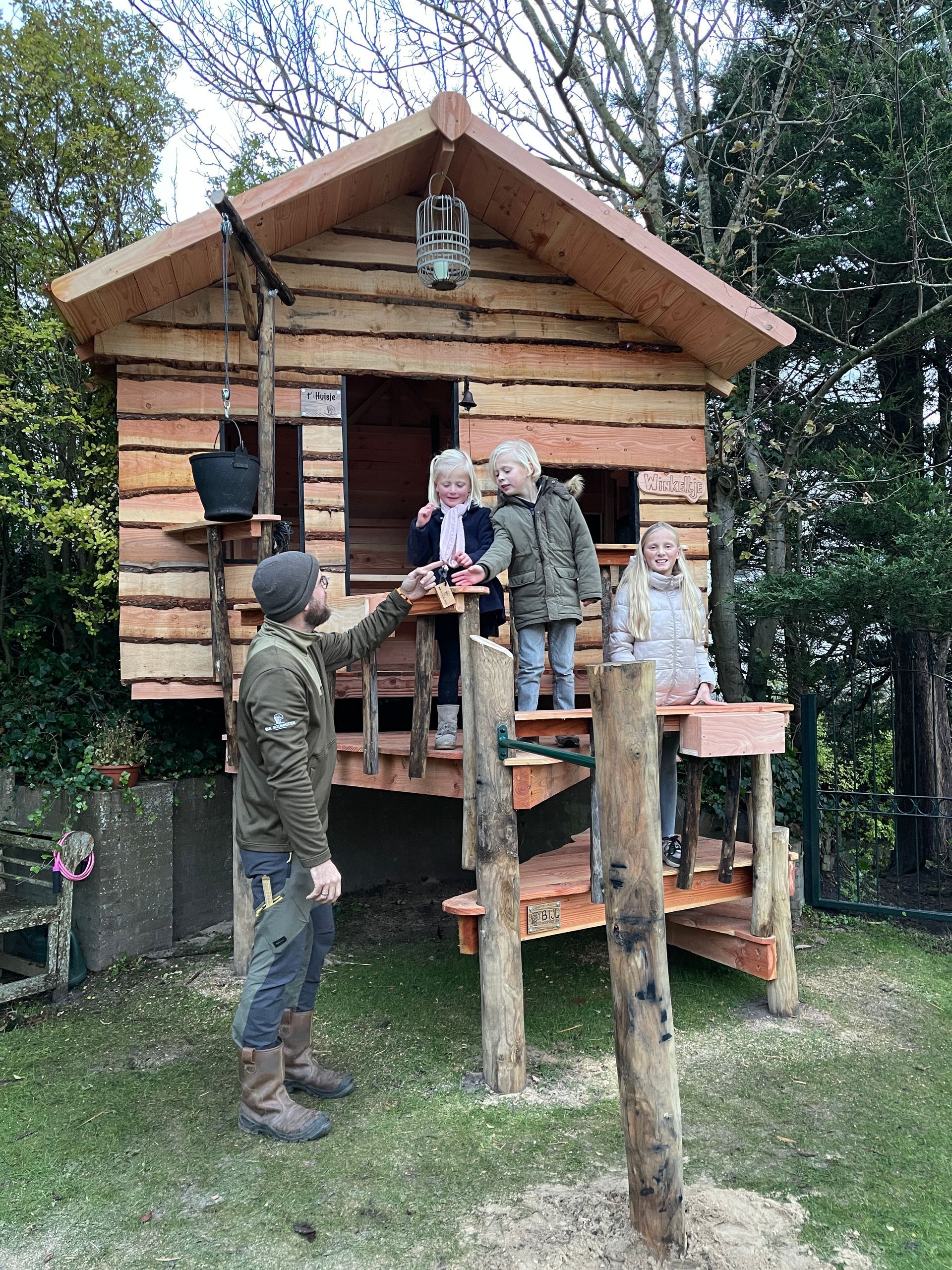 Een man en twee kinderen staan voor een houten boomhut.