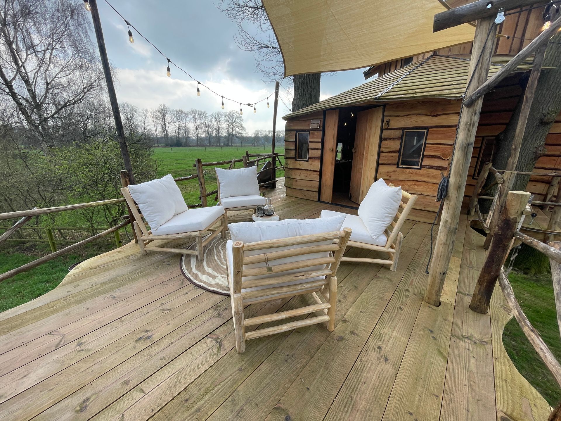 Houten boomhut met terras, zitgelegenheid buiten en sfeerverlichting met uitzicht op een groen veld.