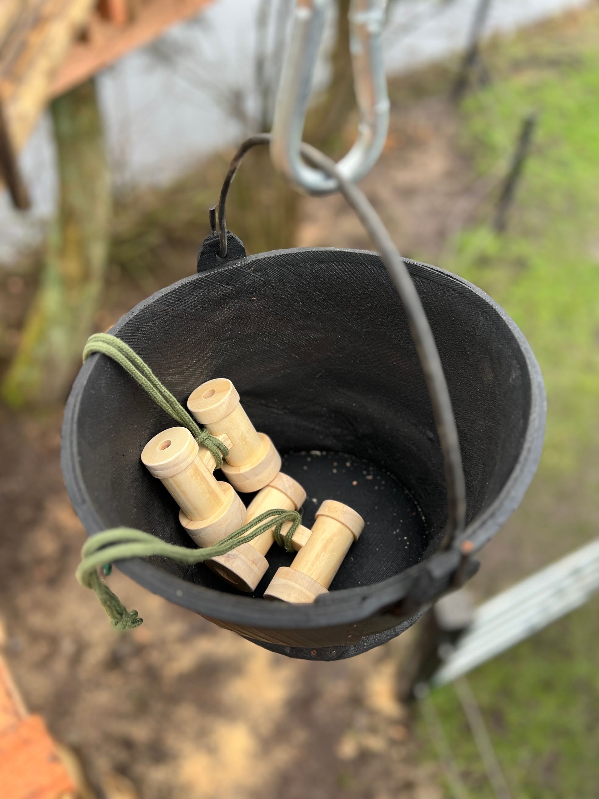 Zwarte emmer met diverse houten stukken erin, buiten opgehangen aan een metalen karabijnhaak.