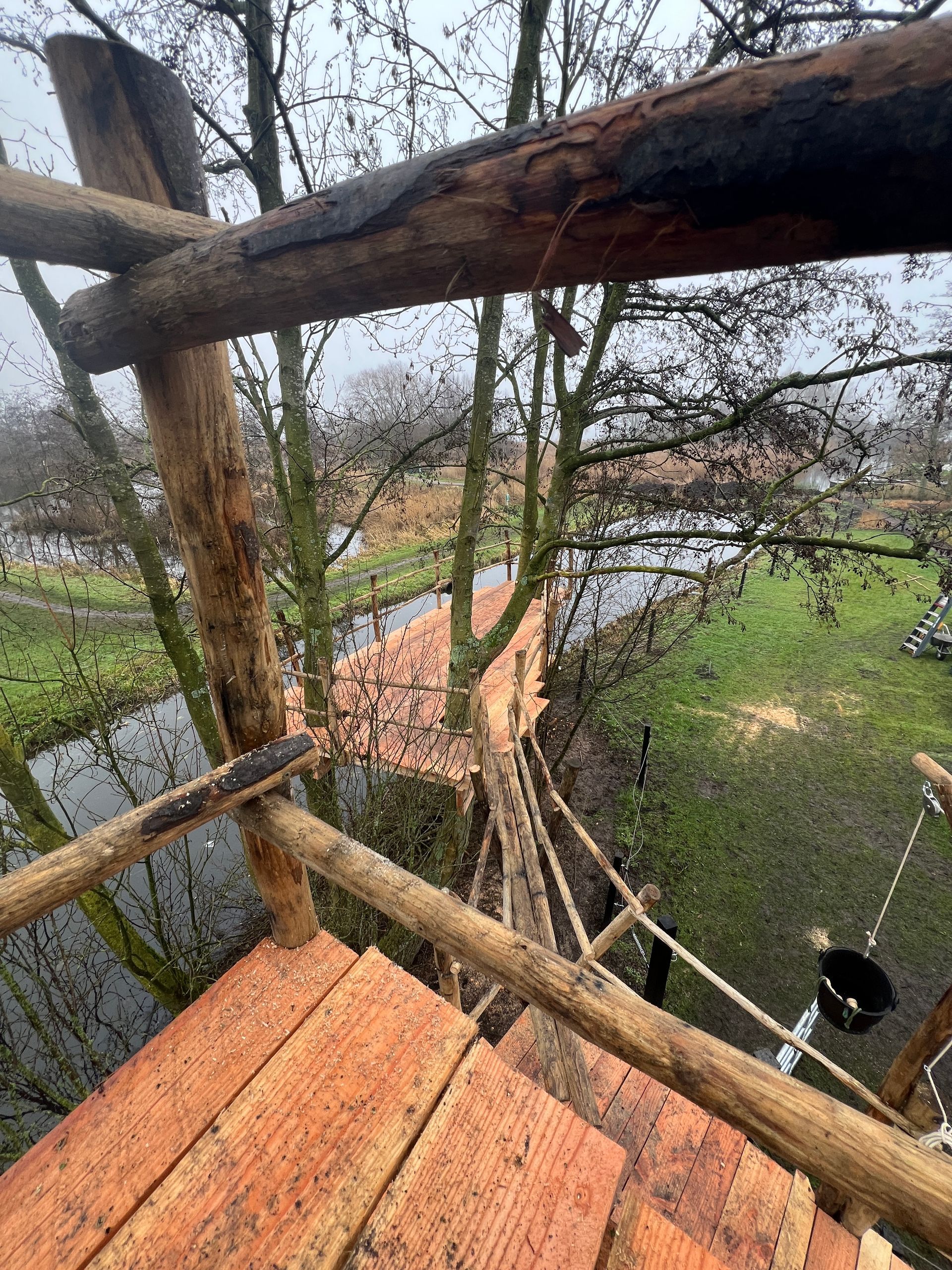 Uitzicht vanaf een platform in een boomhut over een touwbrug, bomen en een waterweg.