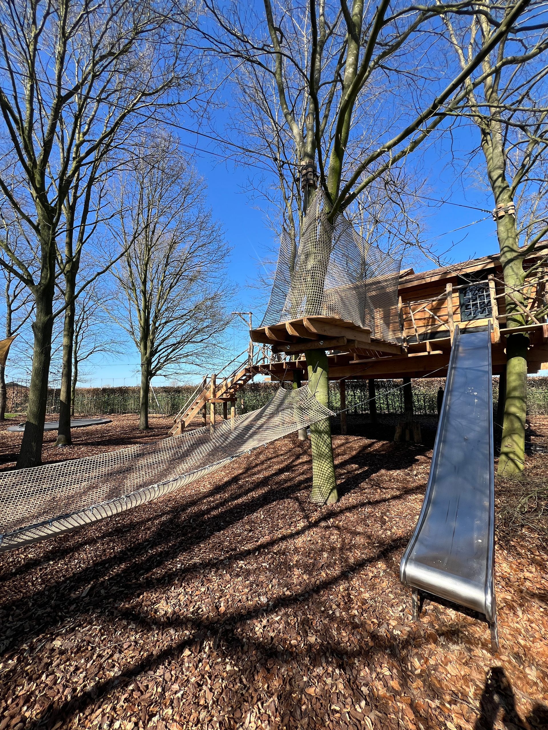 Een houten boomhut met een glijbaan, een brug en een netwerk in een bosrijk gebied onder een blauwe hemel.