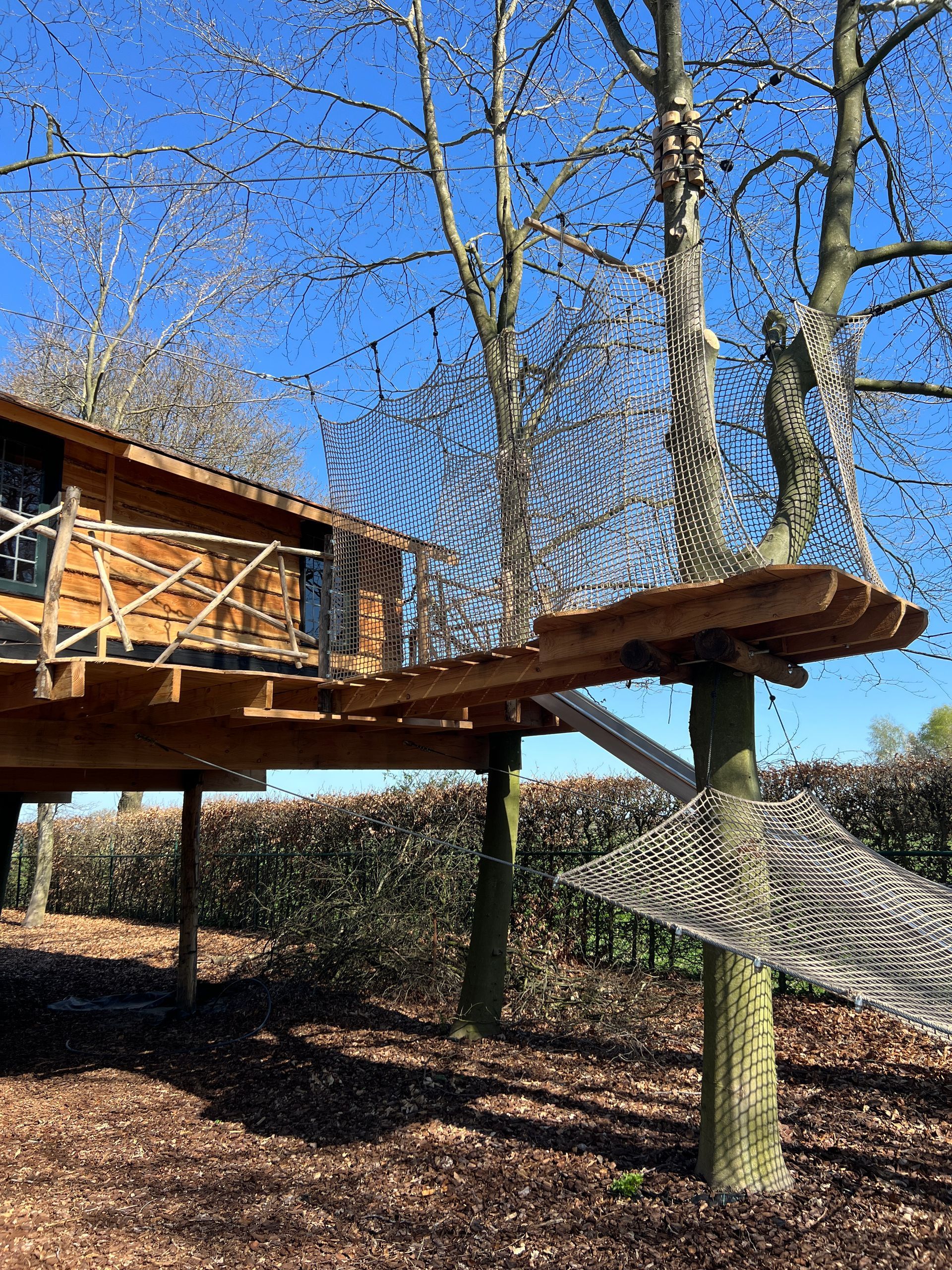 Houten boomhut met vlonder, netten en steunbalken, tegen een blauwe hemel.
