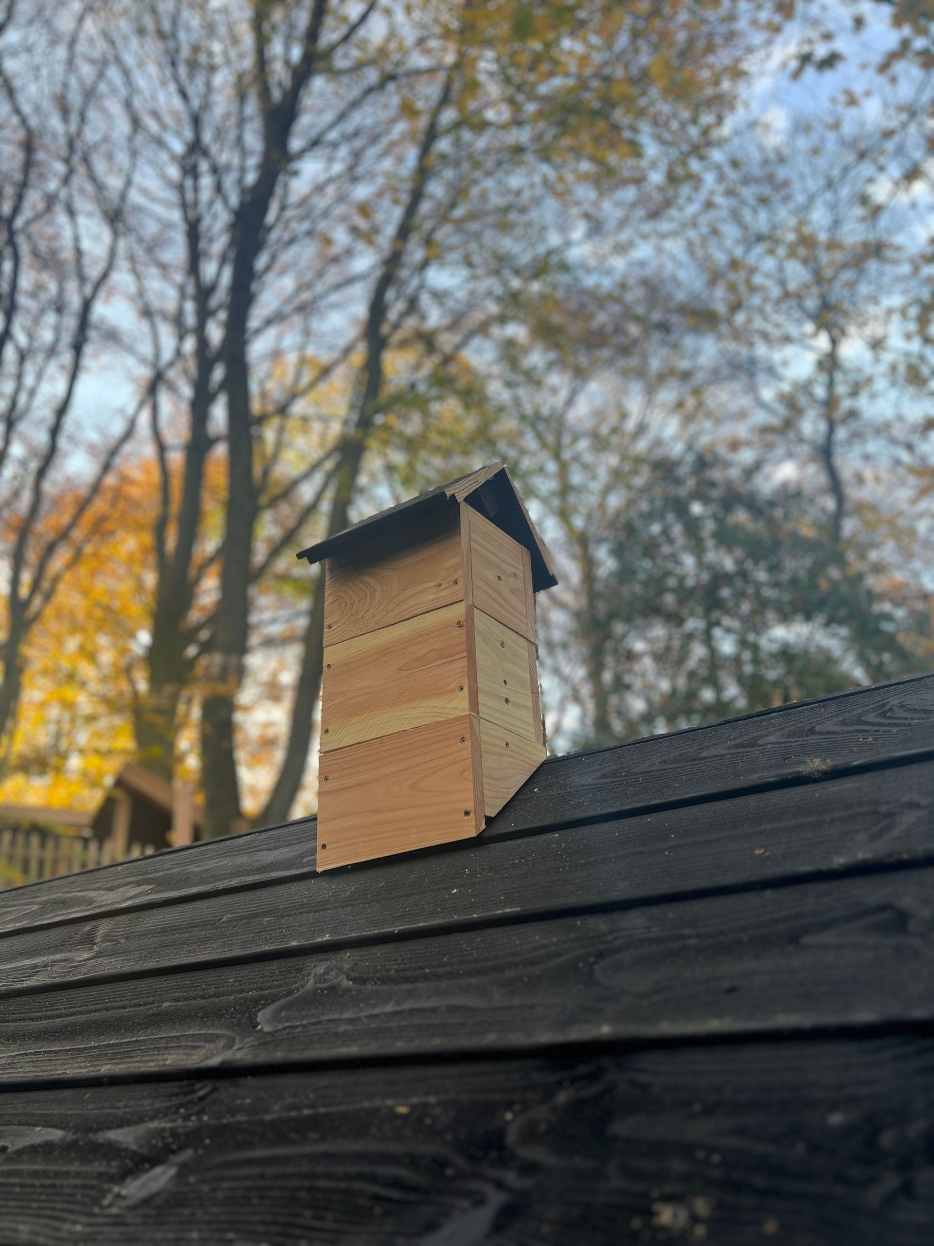 Houten schoorsteen op een donker dak met bomen en een blauwe lucht op de achtergrond.
